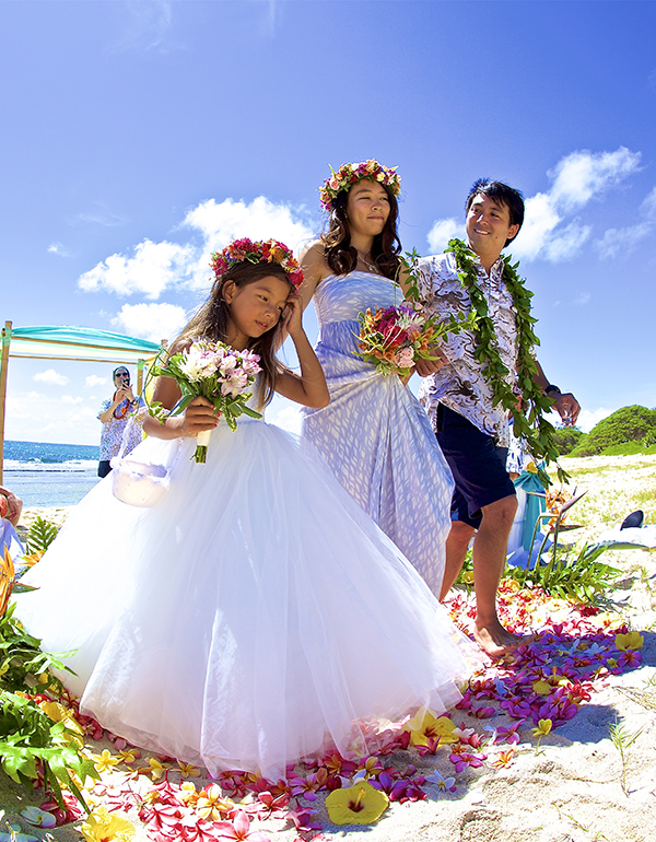 daughter the flower girl walks down the sand flower aise way with her mom and dad after thier Hawaiian style theme wedding
